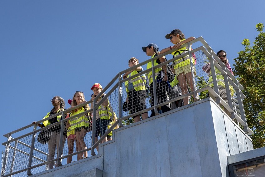 Belvédère en forme de maison des projets avec plusieurs enfants en gilets jaunes de chantier sur la plateforme, devant les palissades du chantier de Noisy-Champs