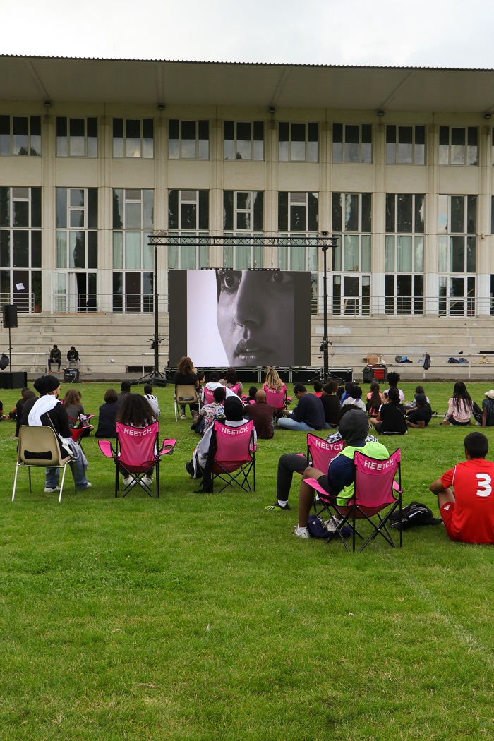 Des spectateurs assis sur des chaises dans un stade de foot avec de la pelouse visionnent le court métrage 0-93. Lab 