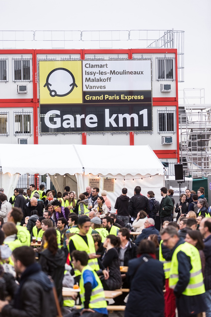 Public en gilet jaune de chantier, de jour, devant le panneau d’entrée du KM1 « Gare KM1 », sur le chantier de la future gare de Fort-d’Issy-Vanves-Clamart-Malakoff