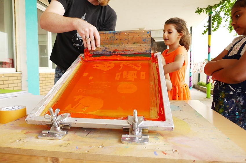Un animateur et deux enfants en train de réaliser une sérigraphie de couleur orange dans le cadre du chantier partagé du collectif « Ne Rougissez Pas »