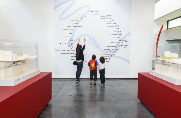 Exposition « Les Passagers du Grand Paris Express », un adulte et deux enfants observent une carte du réseau 
