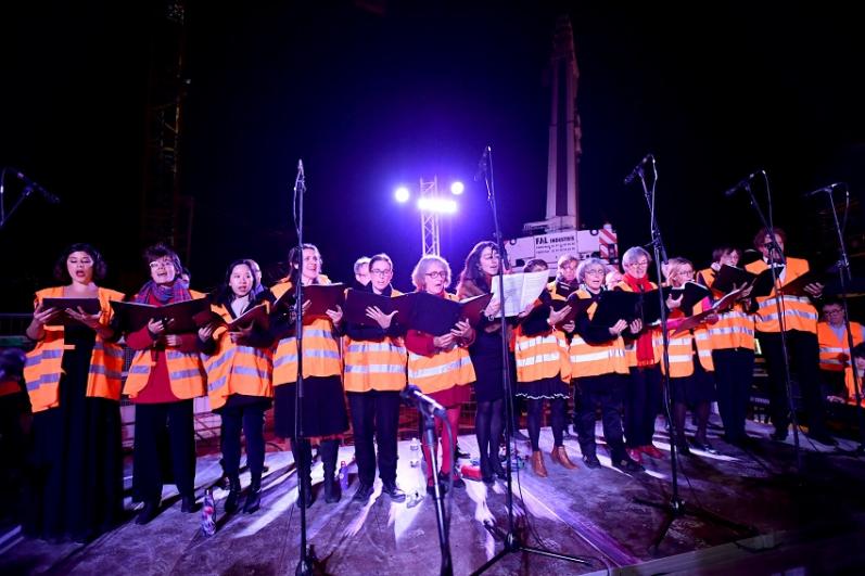 Le chœur de l’Opéra de Massy sur scène de nuit, vêtus de noir et de gilets orange de chantier